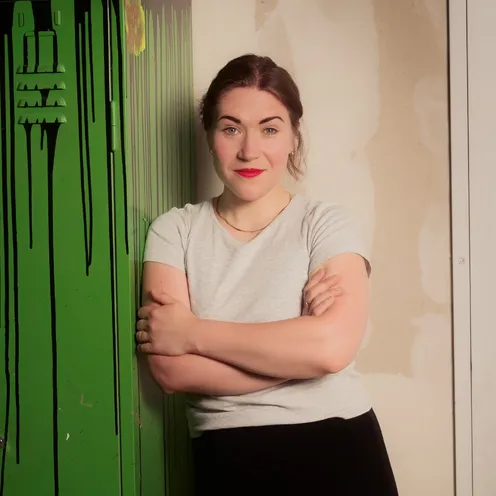 Bildbeschreibung sagte:Eine Frau mit rotem Lippenstift steht mit verschränkten Armen vor einem grünen Spind. Sie trägt ein graues T-Shirt und schwarze Hose, blickt selbstbewusst in die Kamera. Der Hintergrund wirkt industriell mit unbehandelter Wand.