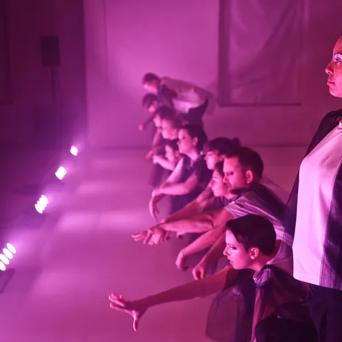 Mehrere Frauen und Männer mit heller bis mittlerer Hautfarbe knien nebeneinander auf einer Bühne im pinken Licht. Ihre Arme sind nach vorn gestreckt, die Gesichter konzentriert. Rechts steht eine Frau mit mittlerer Hautfarbe und lockigem Haar seitlich im Profil.