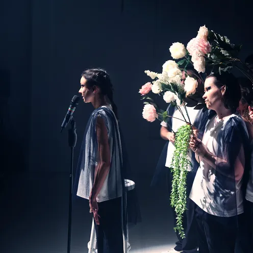 Eine schlanke Frau mit heller Haut steht seitlich an einem Mikrofon im Scheinwerferlicht. Neben ihr halten Frauen und Männer mit heller bis mittlerer Haut große Blumensträuße. Die Gruppe trägt weiße Oberteile mit dunklen, transparenten Stoffen.