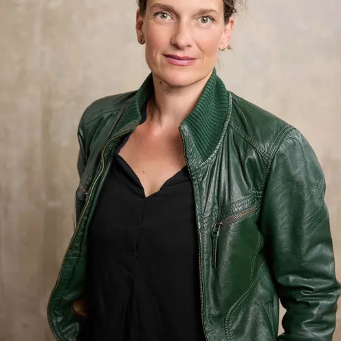 Bildbeschreibung sagte: Eine Frau mit offenem Blick steht vor neutralem Hintergrund. Sie trägt eine grüne Lederjacke über einem schwarzen Oberteil und hat die Hände lässig in den Taschen – ein selbstbewusstes und authentisches Porträt.