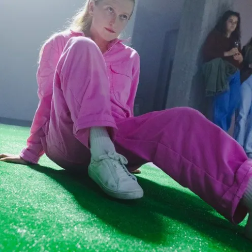 Eine junge Frau in pinkem Overall sitzt lässig auf grünem Kunstrasen. Sie blickt nachdenklich zur Seite, während andere Personen im Hintergrund stehen. Das Licht erzeugt eine kühle, szenische Atmosphäre.