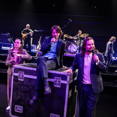 Zwei weiße Frauen und ein weißer Mann mit Mikrofonen performen auf Bühne, eine sitzt auf einem Bühnen-Case, im Hintergrund spielt eine Live-Band.