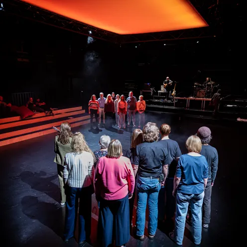 Auf der Bühne steht sich der Bürger:innen-Chor, in zwei Gruppen von je 8-9 Personen geteilt, es sind Personen unterschiedlichen Alters, Geschlecht und Hautfarbe, gegenüber. Im Hintergrund spielt eine Live-Band, auf der Tribüne am linken Rand sitzen ein paar Zuschauer:innen.