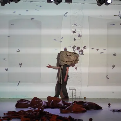 Eine hellhäutige Person steht auf einer Bühne mit Stoff auf dem Kopf, umgeben von projizierten Buchstaben. Auf dem Boden liegen braune Stoffe und Bälle, die Szene wirkt künstlerisch und experimentell.