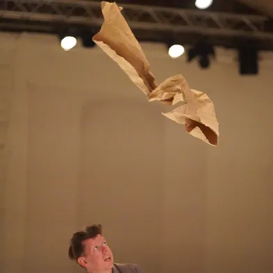 Ein hellhäutiger Mann mit braunen Haaren blickt nach oben auf ein schwebendes Stück braunes Papier. Er trägt ein graues Sakko, die Szene spielt in einem Theaterraum mit Bühnenlicht.
