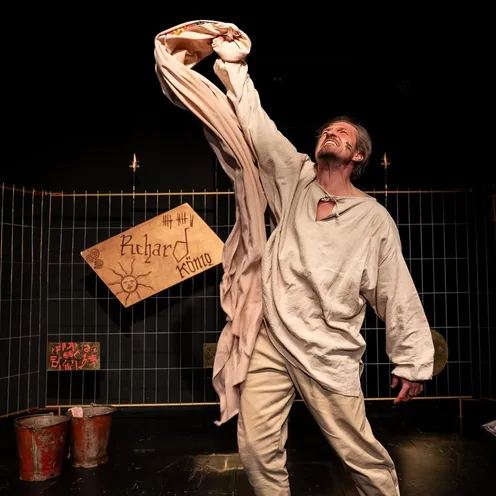 Ein weißer Mann mit Gesichtsbemalung hebt dramatisch ein Tuch in die Luft, trägt ein weißes, mittelalterlich wirkendes Hemd. Er steht auf einer Theaterbühne vor einem Gitter und einem Schild mit der Aufschrift „Richard König“.