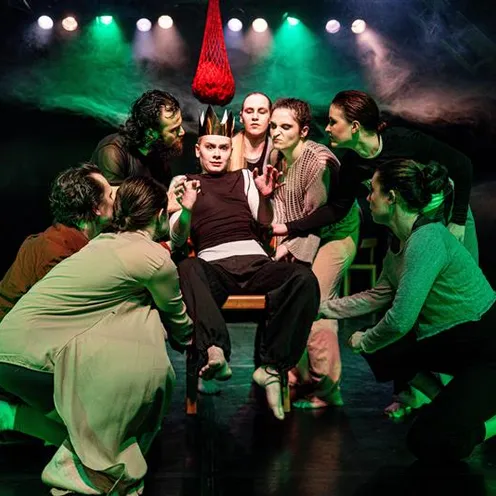 Acht Personen mit heller Haut, vier Frauen und vier Männer, umringen auf einer Bühne einen sitzenden Mann mit heller Haut. Dramatisches grünes Licht, Stühle im Hintergrund, alle barfuß, konzentrierte Gesichter, theatralische Szene mit rotem Netz über ihm.