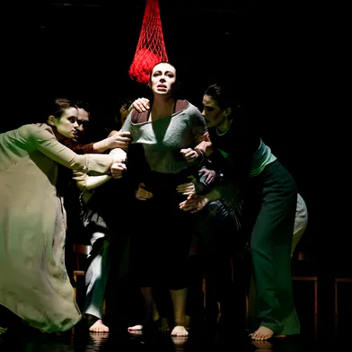 Eine Frau mit heller Haut steht im Mittelpunkt, mehrere Frauen und Männer mit heller Haut halten sie fest. Dunkler Bühnenraum, starkes Licht von oben, rotes Netz über ihr, angespannte Mimik, barfuß, intensive, dramatische Theaterszene.