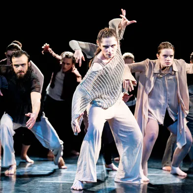Zwei Frauen und mehrere Männer mit heller Haut tanzen auf einer Bühne. Die Frau im Vordergrund steht kraftvoll, die anderen in dynamischen Posen. Schwarzer Hintergrund, barfuß, moderne Kostüme, fokussierte Blicke, energiegeladene Tanzperformance.