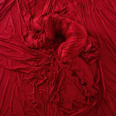 Eine Person liegt eingerollt inmitten von rotem Stoff. Der Körper ist vollständig in das Tuch gehüllt, nur die Form erkennbar. Das Bild wirkt abstrakt, intensiv rot, mit weichen Falten und ruhiger, fast verletzlicher Stimmung.