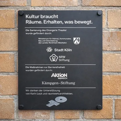 Schwarze Tafel an einer Ziegelwand mit weißem Text: Informationen zur Förderung des Orangerie Theaters Köln durch verschiedene Institutionen. Es sind keine Personen auf dem Bild zu sehen.