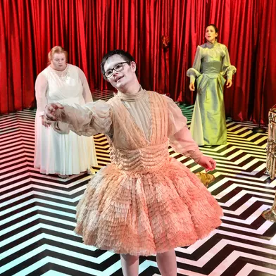 Ein weißer Mann mit Trisomie 21 und Brille tanzt im Vordergrund in einem bauschigen, pfirsichfarbenen Kleid. Zwei weiße Frauen in hellen Kleidern stehen im Hintergrund auf schwarz-weißem Boden.