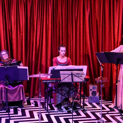 Drei weiße Frauen musizieren vor rotem Vorhang: links mit Akkordeon, Mitte am Keyboard, rechts mit Geige. Alle tragen elegante Kleider in Rosa- und Rottönen, konzentrierter Ausdruck.