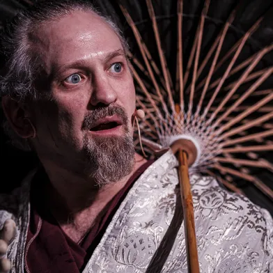 Ein weißer Mann mit heller Haut und grauem Haar blickt überrascht zur Seite. Er trägt einen Bart und Schnurrbart sowie ein historisch wirkendes, hell gemustertes Gewand. Hinter ihm spannt sich ein aufgespannter Schirm auf, dessen Streben strahlenförmig wirken. Dramatisches Licht betont seine blauen Augen.