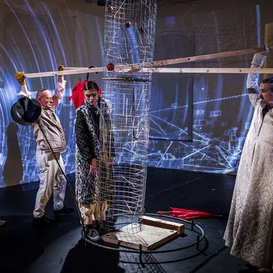 Auf einer Bühne stehen zwei weiße Männer mit heller Haut und eine weiße Frau mit heller Haut. Die Frau ist an eine Konstruktion gebunden, während die Männer Holzbalken halten. Einer trägt einen Hut, der andere ein langes, helles Gewand. Blaues Projektionslicht überzieht den Raum.