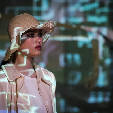 Eine weiße Frau mit heller Haut trägt einen beigen Hut und einen hellen Mantel. Sie blickt ernst zur Seite, während grünlich-blaue Lichtprojektionen über ihr Gesicht und ihre Kleidung verlaufen. Der Hintergrund wirkt unscharf und zeigt abstrakte, technische Muster.