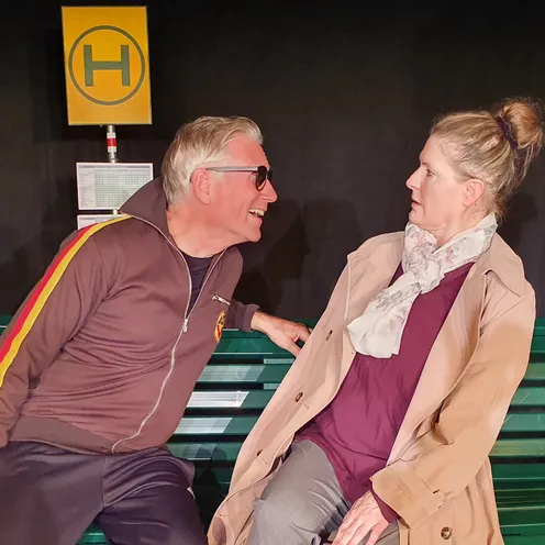 Ein weißer Mann mit heller Haut, grauen Haaren und Sonnenbrille lehnt sich auf einer grünen Bank nach vorn und spricht nah zu einer weißen Frau mit heller Haut und blondem Haar im Dutt. Die Frau trägt einen beigen Mantel und wirkt überrascht. Hinter ihnen steht ein gelbes Haltestellenschild.