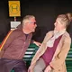 Ein weißer Mann mit heller Haut, grauen Haaren und Sonnenbrille lehnt sich auf einer grünen Bank nach vorn und spricht nah zu einer weißen Frau mit heller Haut und blondem Haar im Dutt. Die Frau trägt einen beigen Mantel und wirkt überrascht. Hinter ihnen steht ein gelbes Haltestellenschild.