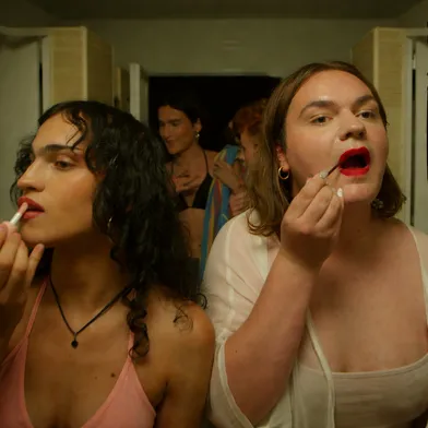 Vier Personen stehen in einem hellen Badezimmer vor Spiegeln und tragen Lippenstift auf. Vorn links eine Frau mit mittlerer, leicht brauner Haut und dunklen lockigen Haaren. Rechts eine Frau mit heller Haut und braunem Haar. Im Hintergrund zwei weitere Frauen mit heller Haut.