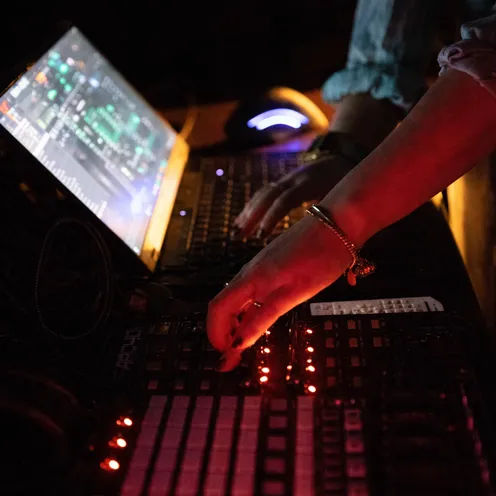 Eine Person, vermutlich eine Frau mit heller Haut, steht in einer dunklen Club- oder Studioumgebung an einem DJ-Mischpult. Ihre Hände bedienen Regler und Fader, daneben leuchtet ein Laptop mit Audioprogramm. Warmes rotes Licht spiegelt sich auf Technik und Armband.