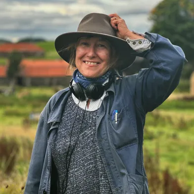 Eine lächelnde Frau mit heller Haut steht draußen auf dem Land. Sie trägt einen breiten Hut, eine blaue Jacke, einen grauen Pullover und Kopfhörer um den Hals. Mit einer Hand hält sie den Hut fest. Im Hintergrund sieht man grüne Felder, Häuser mit roten Dächern und einen bewölkten Himmel.