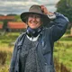 Eine lächelnde Frau mit heller Haut steht draußen auf dem Land. Sie trägt einen breiten Hut, eine blaue Jacke, einen grauen Pullover und Kopfhörer um den Hals. Mit einer Hand hält sie den Hut fest. Im Hintergrund sieht man grüne Felder, Häuser mit roten Dächern und einen bewölkten Himmel.