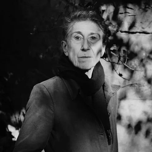 Eine ältere Frau mit heller Haut steht im Freien vor einem dunklen, unscharfen Hintergrund aus Bäumen und Lichtflecken. Das Foto ist schwarz-weiß. Sie trägt einen Mantel und einen dunklen Schal. Ihr kurzer Haarstil und ihr ruhiger, ernster Blick sind deutlich zu erkennen.