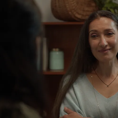 Eine Frau mit heller Haut und langen braunen Haaren steht in einem gemütlichen Raum vor einem Regal mit Pflanzen und Büchern. Sie trägt einen hellblauen Pullover, Ohrringe und eine Kette. Mit verschränkten Armen lächelt sie einer anderen Person gegenüber.