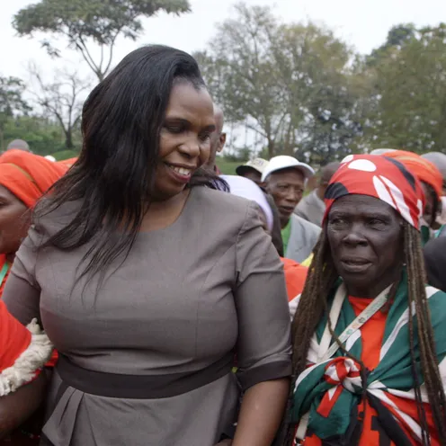 Mehrere Schwarze Frauen mit dunkler Hautfarbe stehen dicht zusammen bei einer Versammlung. In der Mitte lächelt eine Frau mit langen schwarzen Haaren und grauem Kleid. Um sie herum tragen Frauen rote Kleidung, Kopftücher und Fahnenfarben, während sie miteinander sprechen.