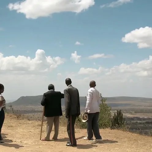 Fünf Menschen stehen auf einem Hügel und schauen in eine weite Landschaft. Zwei Frauen und drei Männer mit dunkler Hautfarbe sind von hinten zu sehen. Einer der Männer stützt sich auf einen Stock. Unter ihnen erstreckt sich eine trockene Ebene unter blauem Himmel mit Wolken.