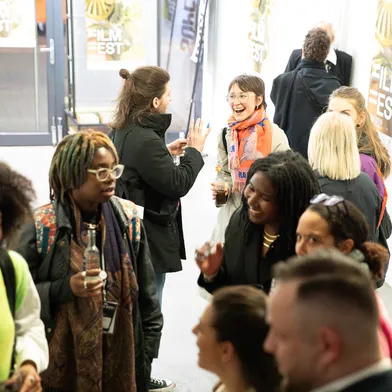 Auf einem Flur bei einem Filmfestival stehen mehrere Menschen in kleinen Gruppen und unterhalten sich. In der Mitte lacht eine Frau mit heller Haut, Brille und orangefarbenem Schal. Um sie herum stehen Männer und Frauen mit heller und dunkler Haut, einige halten Getränke.