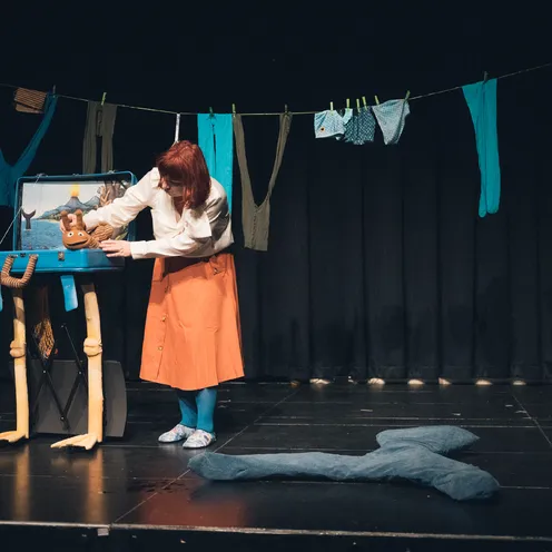 Eine Frau mit heller Haut und roten Haaren beugt sich über einen geöffneten blauen Koffer auf Stelzbeinen auf einer Bühne. Sie trägt eine weiße Bluse, orangefarbenen Rock und blaue Strumpfhose. In dem Koffer steckt eine braune Handpuppe. Über ihr hängt Wäsche an einer Leine.