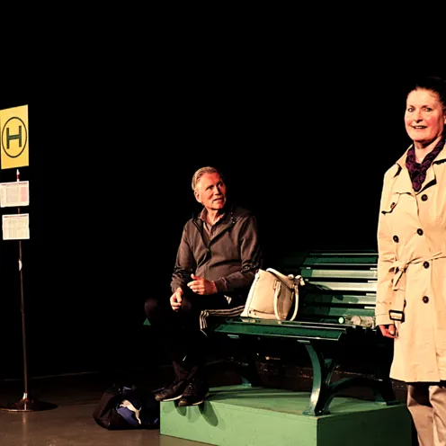 Auf einer dunklen Theaterbühne steht eine inszenierte Bushaltestelle mit grünem Sitz. Ein weißer Mann mit heller Haut und grauen Haaren sitzt auf der Bank und blickt nach rechts. Neben ihm steht eine weiße Frau mit heller Haut und blondem Haar im beigen Mantel. Ein Haltestellenschild steht links.