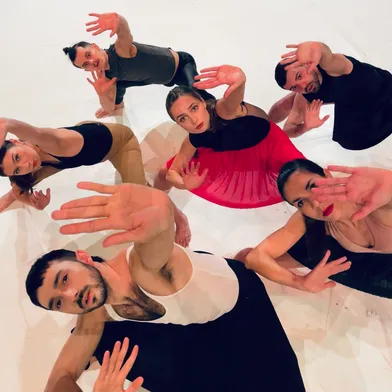 Sechs Personen – drei Frauen und drei Männer mit überwiegend heller bis leicht gebräunter Haut – sind aus der Vogelperspektive zu sehen. Sie tragen Tanzkleidung und strecken ihre Hände zur Kamera. Die Gruppe wirkt dynamisch, die Frau in Rot steht im Zentrum.