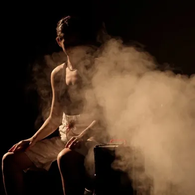 Eine Frau mit heller Haut sitzt leicht nach vorne gebeugt neben einem Metallbehälter, aus dem dichter Dampf aufsteigt. Sie trägt ein dunkles Oberteil und helle Shorts, ihr Gesicht ist teilweise im Schatten verborgen, die Szene wirkt dramatisch.