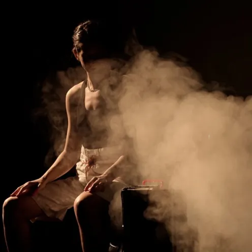 Eine Frau mit heller Haut sitzt leicht nach vorne gebeugt neben einem Metallbehälter, aus dem dichter Dampf aufsteigt. Sie trägt ein dunkles Oberteil und helle Shorts, ihr Gesicht ist teilweise im Schatten verborgen, die Szene wirkt dramatisch.