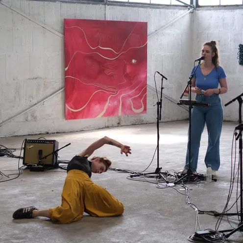 Vier Personen mit heller Haut in einer Industriehalle bei einer Performance. Links spielt eine Frau Gitarre, in der Mitte singt eine Frau am Mikrofon, rechts steht eine weitere Frau an einem Mikrofon. Davor tanzt eine Frau auf dem Boden. Im Hintergrund hängen Kunstwerke.