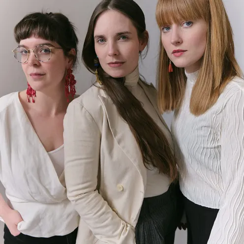 Drei Frauen mit heller Haut posieren vor hellem Hintergrund. Links eine Frau mit Brille und dunklen Haaren, in der Mitte eine Frau mit langen dunklen Haaren und hellem Blazer, rechts eine Frau mit langen roten Haaren. Alle blicken direkt in die Kamera.