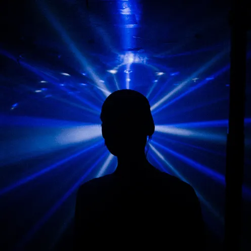 Eine Person steht als schwarze Silhouette vor intensiv blauem Licht. Strahlen breiten sich fächerförmig hinter dem Kopf aus. Geschlecht und Hautfarbe sind wegen der vollständigen Silhouette nicht erkennbar. Die Szene wirkt dunkel und mystisch.