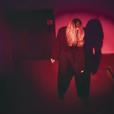 Eine hellhäutige, weiblich wirkende Person mit langen blonden Haaren und schwarzer Kleidung steht gebeugt in einem rot beleuchteten Raum. Das Gesicht wird von den Haaren verdeckt. An der Wand zeichnet sich ein großer dunkler Schatten ab.