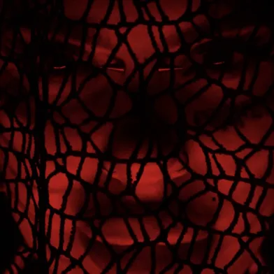 Extreme Nahaufnahme eines Gesichts hinter einem schwarzen, grobmaschigen Netz. Intensives rotes Licht lässt Augen, Nase und Mund schemenhaft hervortreten. Geschlecht und Hautfarbe sind durch Netz, Dunkelheit und farbiges Licht nicht sicher erkennbar.