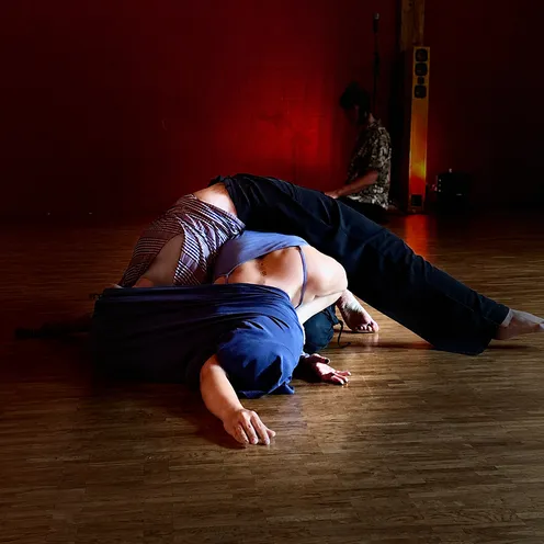 Zwei weiblich wirkende Erwachsene mit heller Haut liegen in einer bodennahen, ineinander verschlungenen Tanzpose. Eine trägt ein blaues Oberteil und schwarze Hose, die andere ein gestreiftes Oberteil. Im Hintergrund sitzt eine weitere Person vor einer roten Wand.