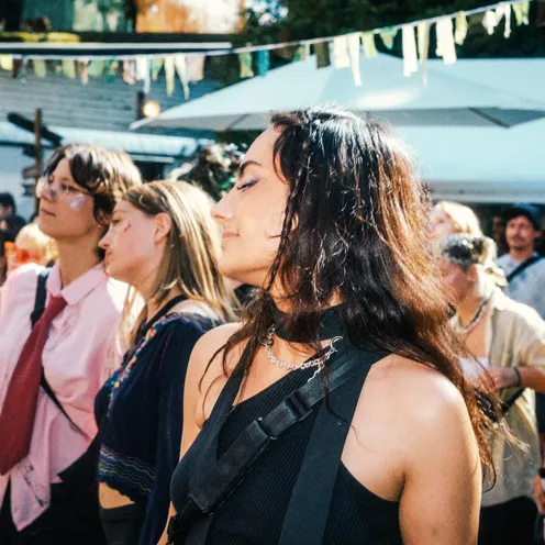 Eine scheinbar junge Frau mit heller bis olivfarbener Haut und langen dunklen Haaren steht mit geschlossenen Augen in einer Menschenmenge auf einem Festival. Im Hintergrund sind weitere Personen unterschiedlicher Hauttöne, Sonnenschirme und Wimpel zu sehen.