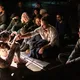 Eine hellhäutige, tätowierte Person sitzt unbekleidet auf dem Boden eines dunklen Raums. Ein hellhäutiger Mann hält ihre Hand, während Frauen und Männer unterschiedlichen Alters dicht um die Szene sitzen und zusehen.