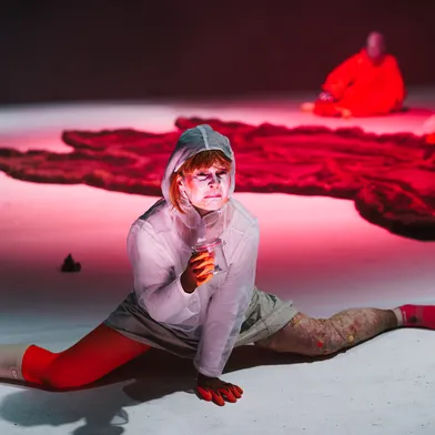 Eine hellhäutige Frau sitzt im Spagat auf einer Theaterbühne und hält ein Glas. Sie trägt helle Kleidung, rote Strümpfe und auffälliges Make-up. Im rot beleuchteten Hintergrund sitzen zwei weitere Frauen neben einer dunklen Plane.