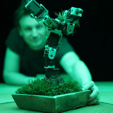 Ein mechanischer Roboter mit zwei erhobenen Armen steht auf einer mit Kunstrasen gefüllten Schale. Dahinter beobachtet eine hellhäutige Frau mit dunklem Haar das Objekt. Grünes Licht prägt die dunkle, futuristische Szene.