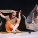 Eine hellhäutige Frau mit roten Haaren sitzt unter einer großen Plane, trägt beige Kleidung, orange Strumpfhose und weiße Stiefel. Hinter ihr sitzt eine dunkelhäutige Frau in heller Kleidung vor einer zeltartigen Plane.