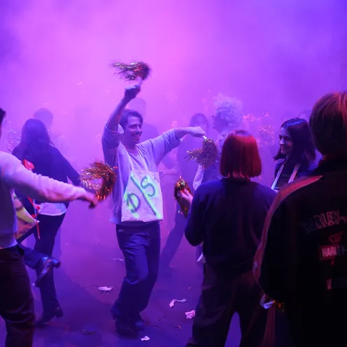 In violettem Licht und dichtem Nebel tanzen mehrere überwiegend hellhäutige Frauen und Männer gemeinsam. Einige schwenken glitzernde Pompons. Im Mittelpunkt hebt ein Mann lachend die Arme, während sich die Gruppe um ihn bewegt.