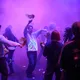 In violettem Licht und dichtem Nebel tanzen mehrere überwiegend hellhäutige Frauen und Männer gemeinsam. Einige schwenken glitzernde Pompons. Im Mittelpunkt hebt ein Mann lachend die Arme, während sich die Gruppe um ihn bewegt.