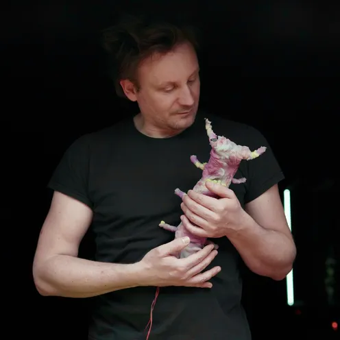 Ein hellhäutiger Mann mit dunklem Haar und schwarzem T-Shirt hält eine kleine, rosa-weiße, organisch wirkende Figur in den Händen und betrachtet sie. Größere ähnliche Formen hängen seitlich im Vordergrund; Lichtstäbe leuchten dahinter.